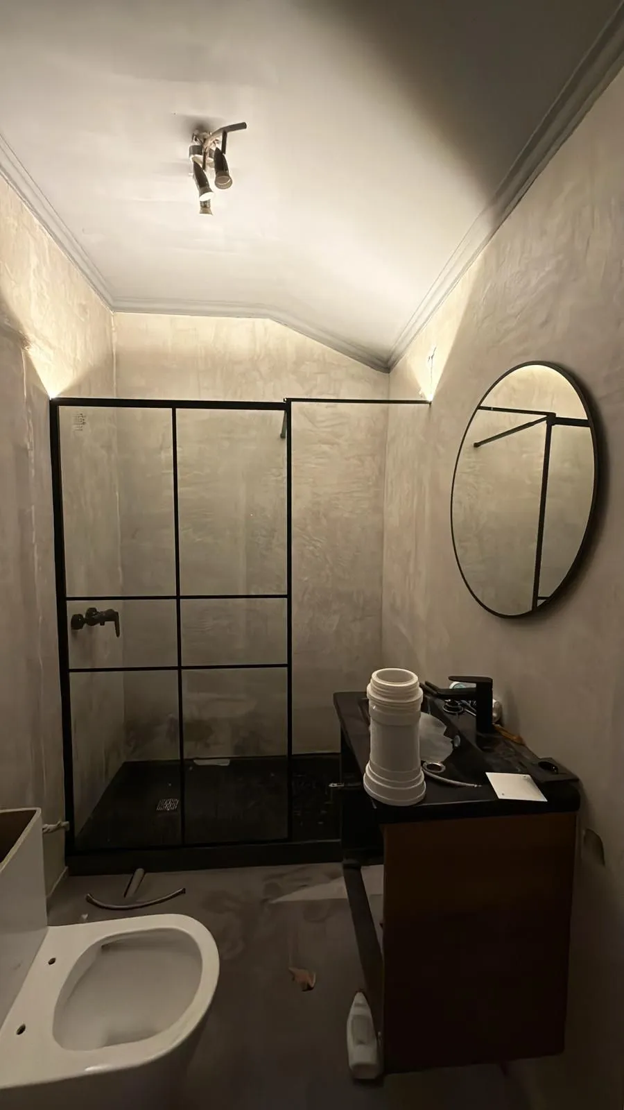 A mesma casa de banho na reta final da obra: microcimento aplicado, duche com vidro de perfil preto e espelho redondo, ainda com material de canalização à vista